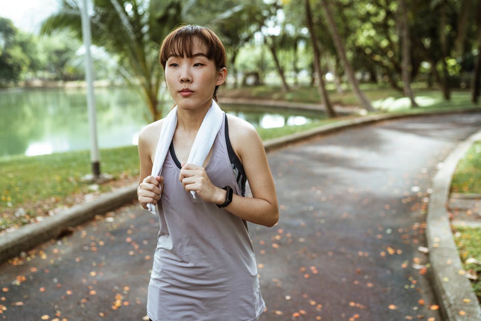 An athletic woman jogging outdoors in a park with a towel around her neck, enhancing wellness.