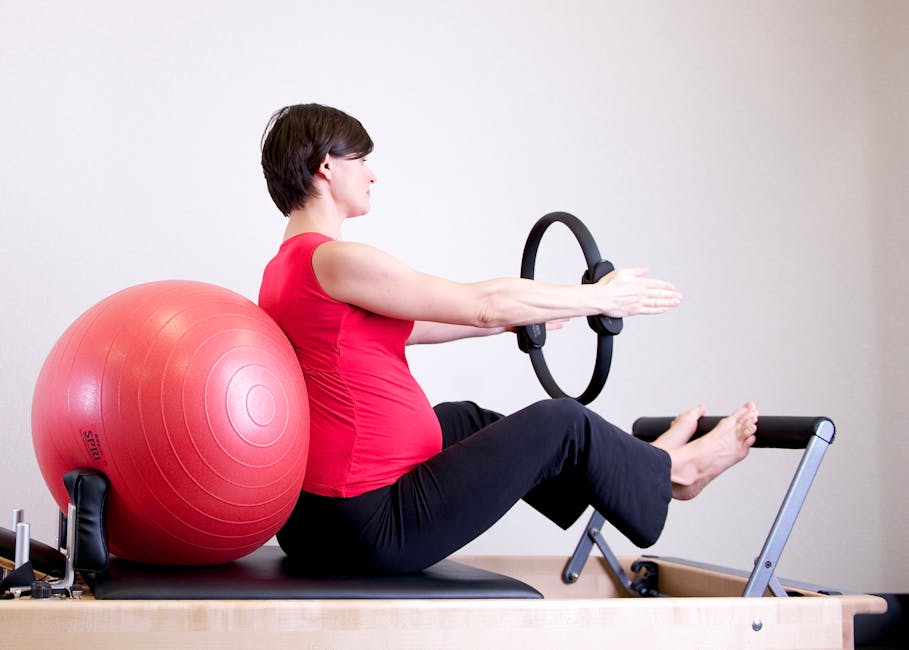 A pregnant woman in a red top practicing Pilates using a fitness ring and exercise ball in a gym setting